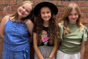 Three little girls stand in front of red brick